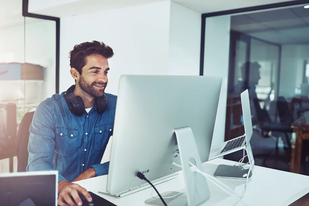 Ein junger Mann mit Kopfhörern um den Hals arbeitet an einem großen Monitor in einem modernen Büro mit Glaswänden und mehreren Bildschirmen.