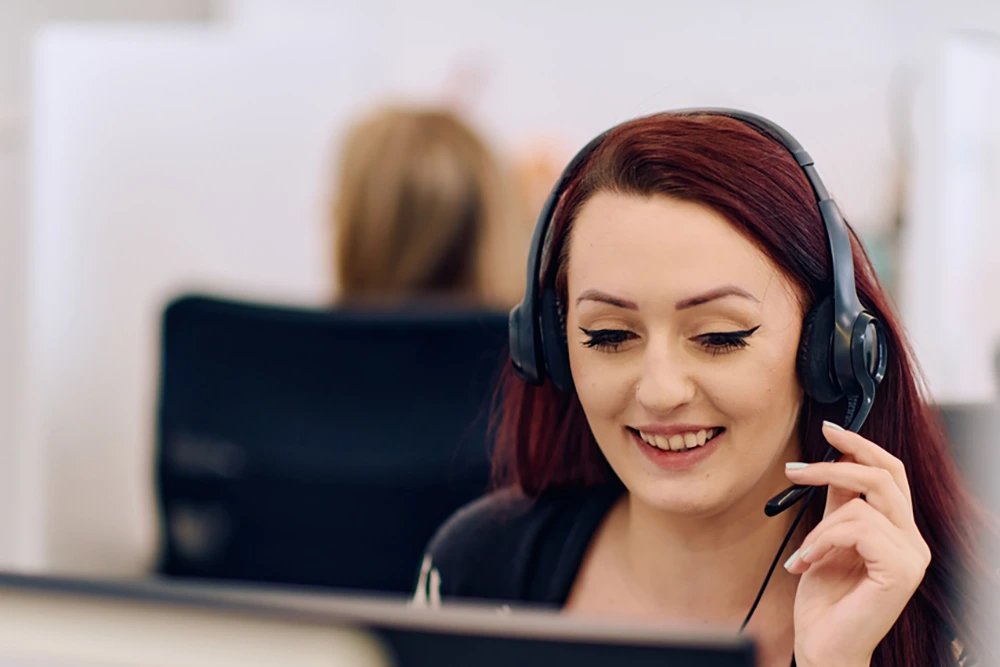 Eine Frau mit dunklen Haaren und Headset lächelt während eines Kundengesprächs. Sie befindet sich in einem modernen Büro mit Arbeitsplätzen im Hintergrund.