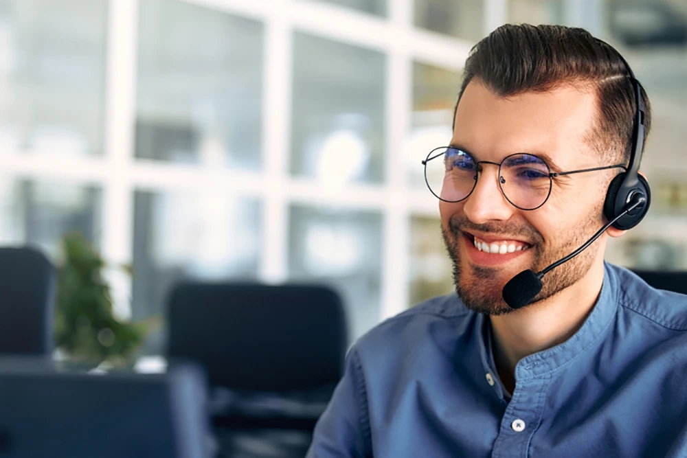 Ein junger Mann mit Brille und Headset lächelt während eines Gesprächs am Arbeitsplatz in einem modernen Großraumbüro.