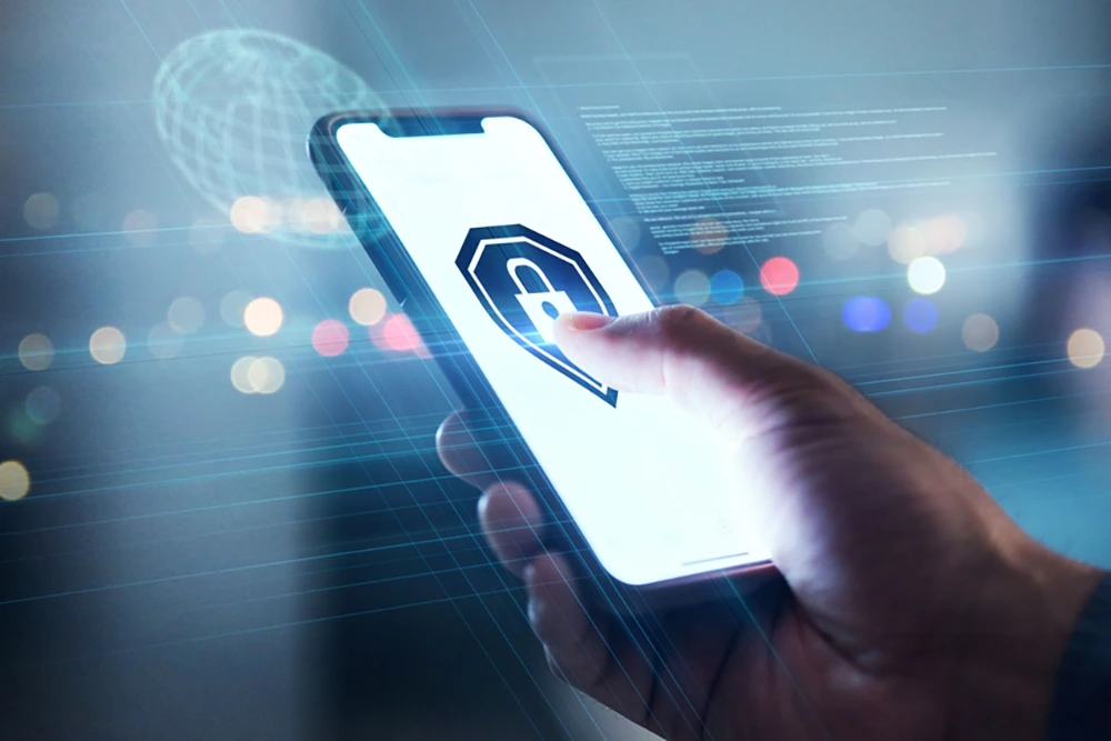 Eine Hand hält ein Smartphone mit einem großen Datenschutzsymbol auf dem Display. Im Hintergrund sind Lichtpunkte und digitale Interface-Elemente sichtbar, was auf IT-Sicherheit hinweist.
