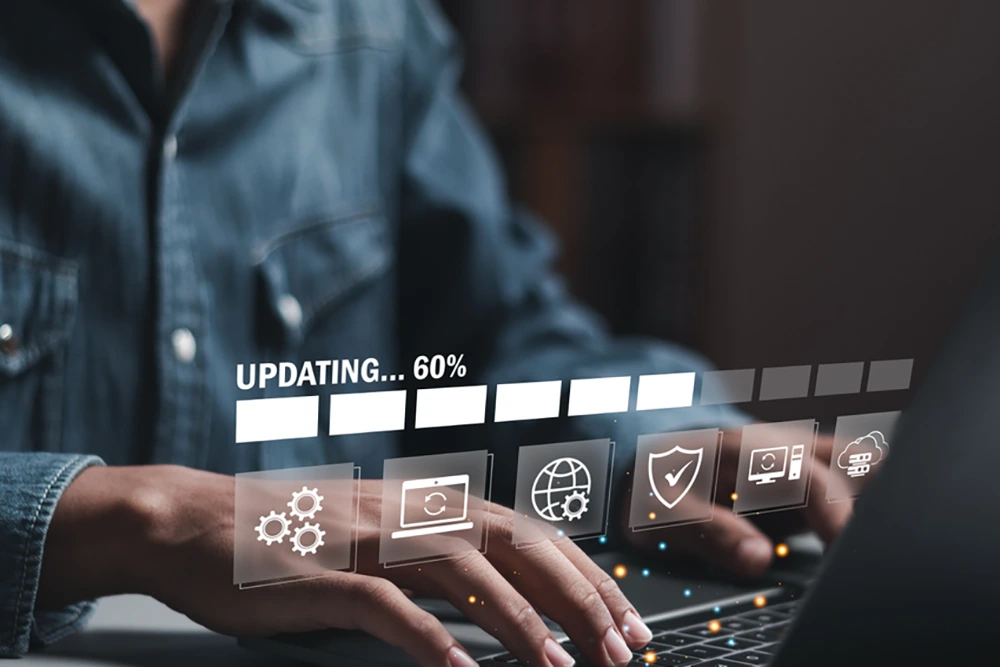 Eine Person tippt auf einem Laptop, über dem eine Fortschrittsanzeige mit der Aufschrift „Updating… 60%“ sowie Symbole für Systemeinstellungen, Schutz und Netzwerkeingabe eingeblendet sind.