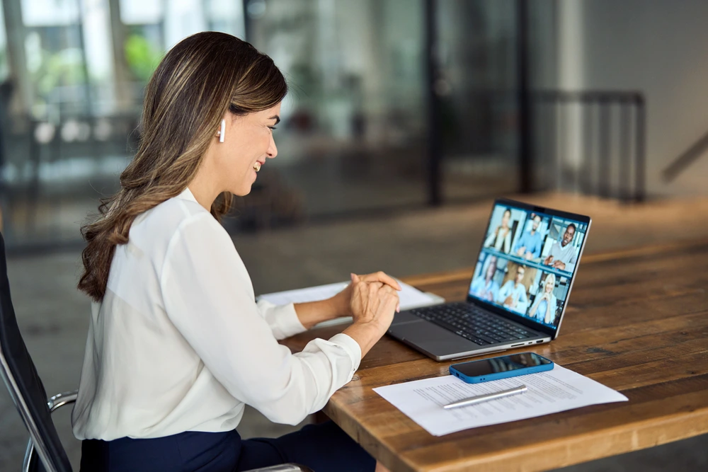 Eine Frau mit weißen Ohrhörern sitzt an einem Holztisch und nimmt an einer Videokonferenz auf einem Laptop teil. Auf dem Bildschirm sind mehrere Gesprächsteilnehmende sichtbar, daneben liegen Unterlagen und ein Smartphone.