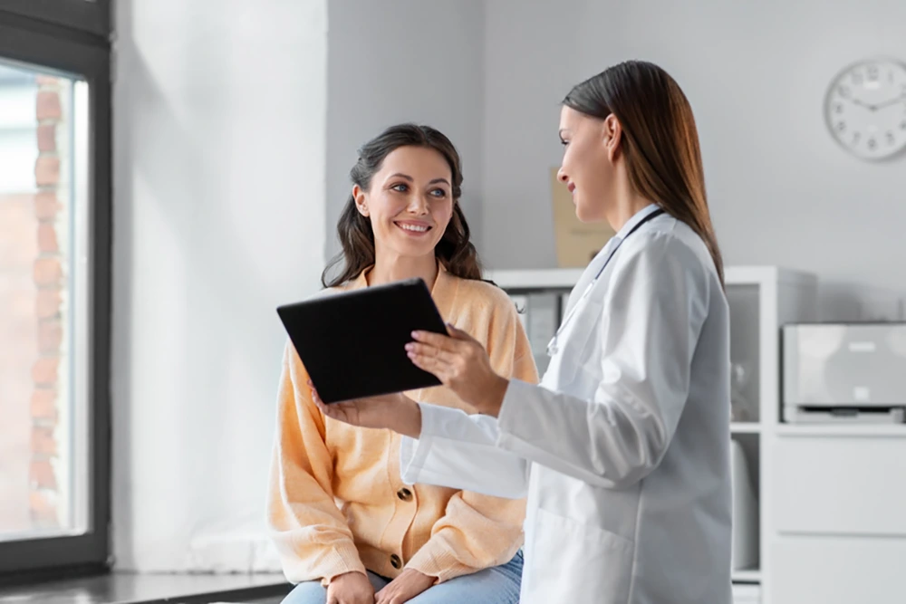 Eine Ärztin zeigt einer lächelnden Patientin etwas auf einem Tablet. Die beiden sitzen in einer hellen Arztpraxis mit großen Fenstern und Regalen im Hintergrund.