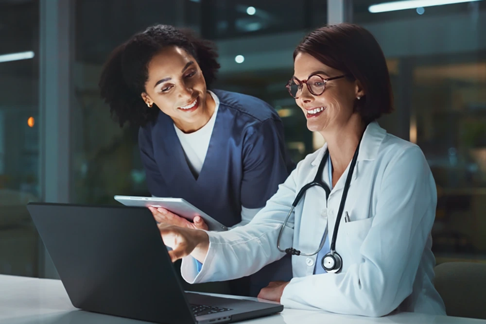 Zwei Ärztinnen in heller Kleidung mit Stethoskopen sitzen an einem Laptop und sehen gemeinsam auf den Bildschirm, eine von ihnen hält zusätzlich ein Tablet.