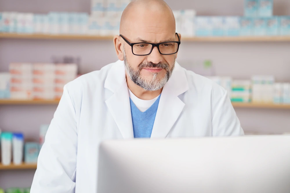 Ein älterer Mann im weißen Kittel und mit Brille arbeitet an einem Computer in einer Apotheke. Im Hintergrund sind Regale mit Medikamentenverpackungen sichtbar.