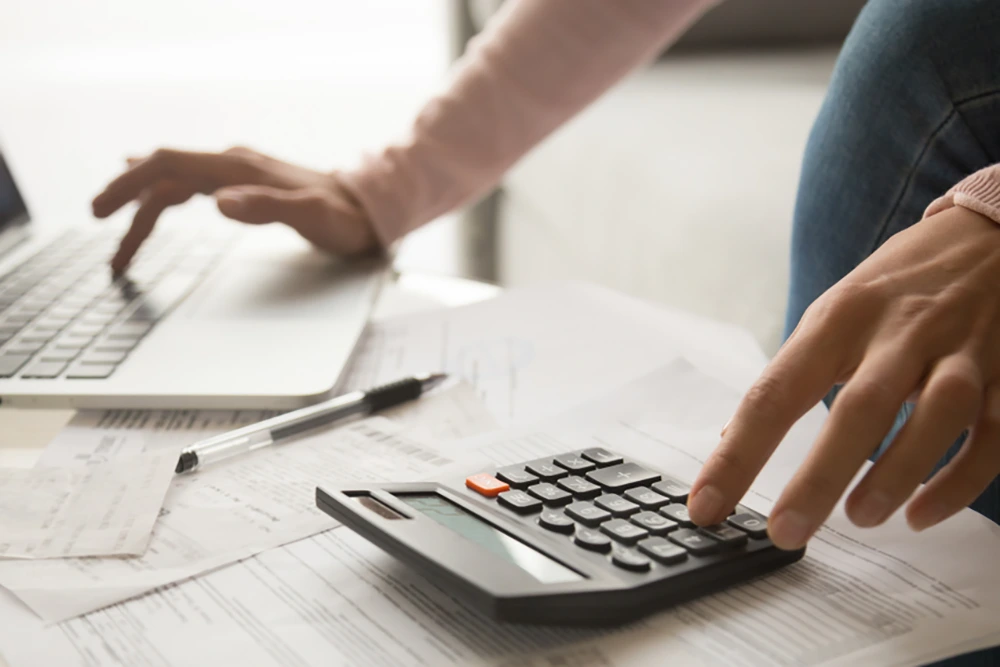 Zwei Hände arbeiten an einem Schreibtisch: Eine benutzt einen Taschenrechner, die andere einen Laptop. Auf dem Tisch liegen Rechnungen, Belege und ein Kugelschreiber.