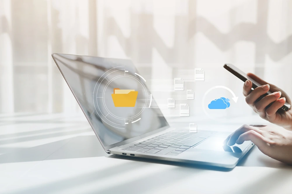 Eine Hand bedient ein Smartphone, während die andere auf der Tastatur eines Laptops liegt. Über dem Bildschirm schweben digitale Symbole für Cloud-Speicherung und Dateizugriff.