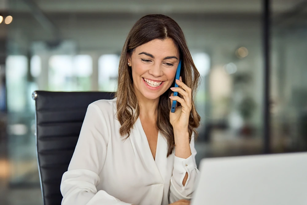 Eine Frau in weißer Bluse telefoniert mit dem Smartphone und lächelt dabei. Sie sitzt an einem Schreibtisch mit geöffnetem Laptop in einem modernen Büro.