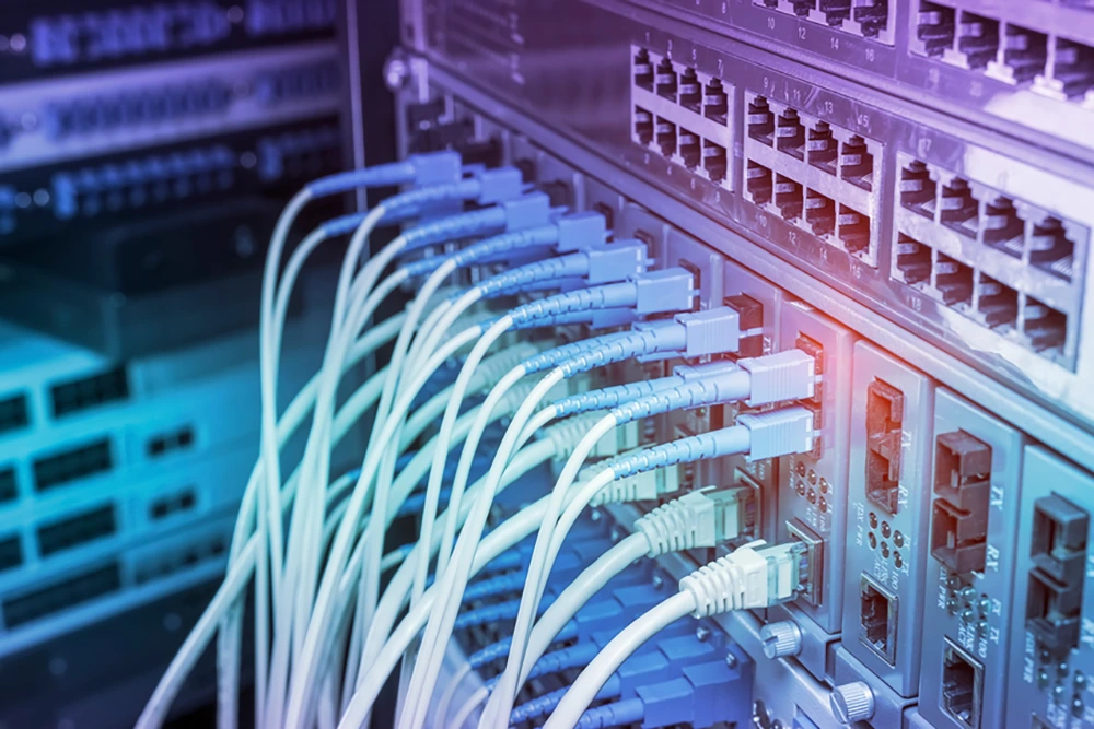 Nahaufnahme eines Serverracks mit zahlreichen eingesteckten Netzwerkkabeln und Anschlüssen. Die Ausleuchtung ist in Blau- und Violetttönen gehalten.